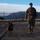 A military service member in full camouflage uniform and tactical vest walks with a military working dog (appears to be a Belgian Malinois) on a leash across a paved surface. The setting appears to be a military installation, evidenced by the chain-link fence with barbed wire in the background and arid, mountainous terrain. The soldier is wearing a black beret and smiling toward the camera.