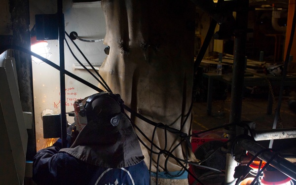 A worker wearing a welding helmet and dark coveralls with a trident/anchor logo on the back is performing welding or grinding work in a confined industrial space, likely aboard a ship or in a shipyard. Sparks are visible near the work area, and the space is illuminated by portable work lights. Multiple cables and hoses are visible throughout the cluttered workspace.