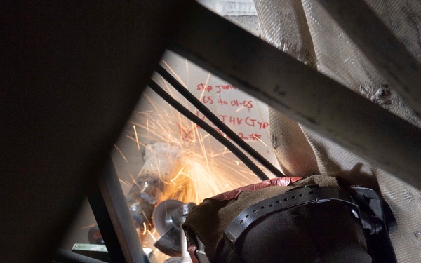 A welder in full protective gear is performing welding or cutting work in a confined, industrial space, with visible sparks flying from the work area. The worker is wearing a welding helmet, leather gloves, and a fire-resistant jacket, and appears to be working through a structural opening. Red text markings are visible on a surface in the background, likely indicating engineering or construction specifications.