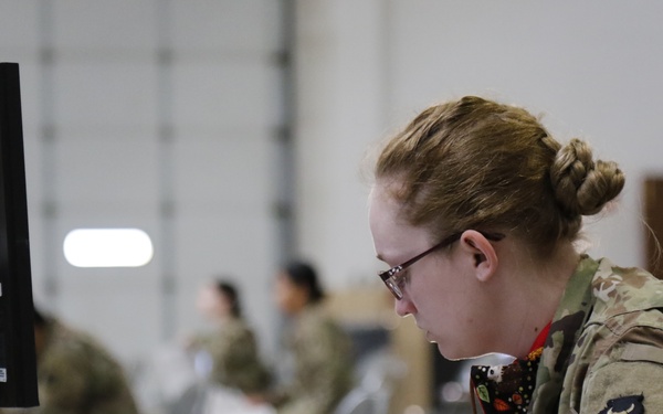 A female military service member in OCP (Operational Camouflage Pattern) uniform is working at a computer workstation in what appears to be a large indoor military facility or hangar. She is leaning forward intently, reviewing documents while operating a computer. Multiple other uniformed personnel are visible in the blurred background, suggesting a large-scale administrative or processing operation.