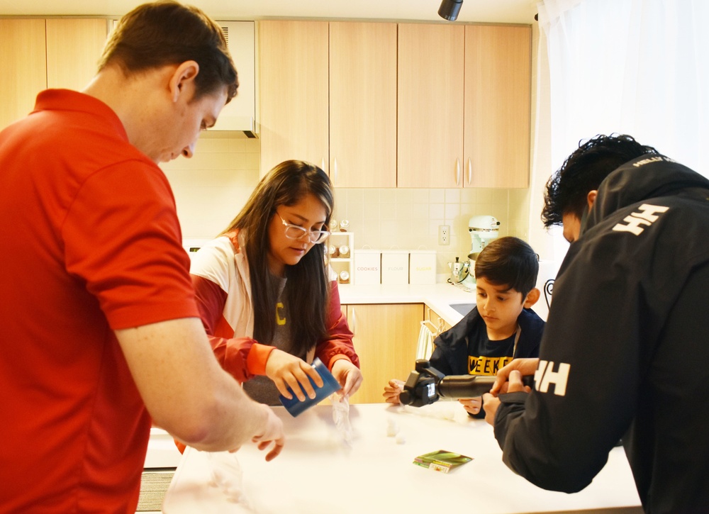 Four people — one adult and three younger individuals — are gathered around a kitchen counter engaged in a hands-on activity. The scene appears to take place in a modern, well-lit kitchen with light wood cabinetry. They appear to be working with materials that may involve dry ice or a similar substance, given the visible vapor/smoke on the counter.