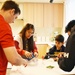 Four people — one adult and three younger individuals — are gathered around a kitchen counter engaged in a hands-on activity. The scene appears to take place in a modern, well-lit kitchen with light wood cabinetry. They appear to be working with materials that may involve dry ice or a similar substance, given the visible vapor/smoke on the counter.