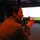 A female soldier in camouflage uniform is aiming a rifle toward a large projection screen in what appears to be an indoor weapons training simulator. She is in a crouched or kneeling position, holding the weapon in a firing stance. The environment is dimly lit with a reddish ambient light, consistent with a military simulation training facility.