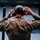 A person wearing U.S. Marine Corps MARPAT digital camouflage uniform is photographed from behind, adjusting tactical hearing protection earmuffs on their head. Their dark hair is worn in a bun, and they appear to be at an indoor shooting range. The image captures a preparatory moment before a firearms training exercise.