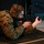 A young male in U.S. Marine Corps MARPAT digital camouflage uniform is in a prone shooting position on a black mat, aiming a rifle downrange. He is wearing tactical hearing protection (ear muffs) and appears to be at an indoor shooting range. The setting suggests a training or demonstration environment.