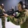 A female soldier wearing Australian Army camouflage uniform is handling an M16/AR-15 style rifle at what appears to be an indoor shooting range or training facility. She is wearing tactical hearing protection (earmuffs) and has a patch on her sleeve reading 'AUSTRALIA'. Other military personnel in camouflage uniforms are visible in the background.