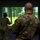 Two military personnel are shown from behind inside what appears to be a simulation training facility. A large projected screen displays a tropical or subtropical outdoor environment with trees and green vegetation. One individual in MARPAT digital camouflage (likely US Marine Corps) observes while another in a different camouflage pattern appears to be actively engaged with the simulation.