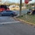 A vehicle accident scene showing a gray sedan that has crashed near a street corner, with debris scattered across the road and curb area. Fire Rescue Engine E-1 is present on the left side of the scene. Emergency responders and bystanders are attending to what appears to be a victim on the ground near the fence of St. Michael and All Angels Episcopal Church.