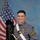 A young man in a United States Military Academy (West Point) cadet dress gray uniform poses for a formal portrait photograph. He is holding a full-dress tar bucket hat with black feather plume and a ceremonial saber, with an American flag visible in the background. His uniform displays sergeant rank chevrons, a medal, airborne wings, and gold buttons consistent with West Point cadet regalia.