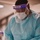 A healthcare worker is dressed in full personal protective equipment (PPE) in what appears to be a medical facility. The individual is handling what appear to be nasal swabs, suggesting preparation for or completion of COVID-19 or similar diagnostic testing. The image captures a close-up, portrait-oriented view with the worker leaning slightly forward.