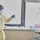 A healthcare worker in full personal protective equipment (PPE) is working in what appears to be a hospital patient room or ICU setting. The worker is handling medical tubing or a line while standing near IV infusion pumps and medical equipment. The setting suggests an isolation or high-acuity care environment.