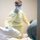 A healthcare worker wearing full personal protective equipment (PPE) is attending to a patient in what appears to be a hospital or clinical setting. The worker is dressed in a yellow isolation gown, blue gloves, surgical mask, and a labeled face shield. A second person, partially visible on the right, appears to also be present near the patient.