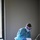 A healthcare worker in full personal protective equipment (PPE) is attending to a patient lying in a hospital bed. The worker is leaning over the patient and appears to be reviewing or presenting a document. The setting is a dimly lit hospital room with a window visible on the left side.