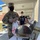 A U.S. Army soldier in OCP (Operational Camouflage Pattern) uniform and black face mask is serving or distributing food from a chafing dish on a table outdoors under a covered walkway. The soldier is wearing latex gloves and holding a plastic 'Thank You' grocery bag, suggesting a food distribution or meal service event. Several other masked individuals are visible in the background waiting in line.