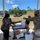 A U.S. Army soldier in OCP camouflage uniform and black face mask is handing a plastic bag of food to a civilian woman wearing a face mask and gloves at an outdoor food distribution event. Multiple people are present under a covered pavilion, with a cart holding food items including what appear to be oranges. The setting appears to be a military installation or community facility, consistent with COVID-19 era food assistance operations.