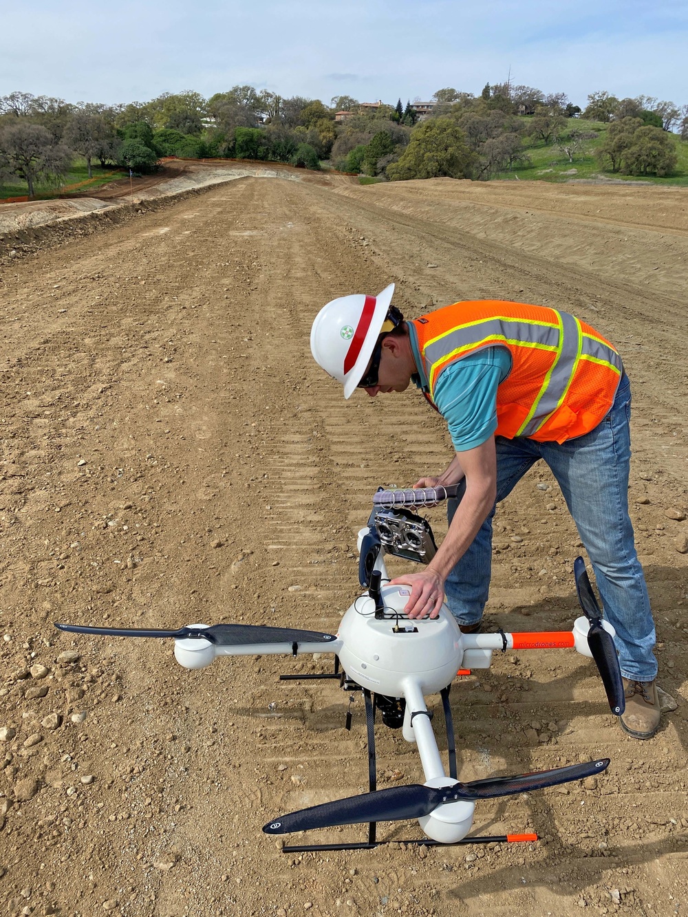 A construction worker in safety gear is preparing or inspecting a professional-grade quadcopter drone on a graded dirt construction site. The site appears to be a large earthwork or road construction project, with compacted soil extending toward a tree-lined hillside with residential structures in the background. The drone is equipped with a multi-lens camera payload, suggesting it is being used for aerial surveying or mapping.