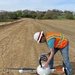 A construction worker in safety gear is preparing or inspecting a professional-grade quadcopter drone on a graded dirt construction site. The site appears to be a large earthwork or road construction project, with compacted soil extending toward a tree-lined hillside with residential structures in the background. The drone is equipped with a multi-lens camera payload, suggesting it is being used for aerial surveying or mapping.