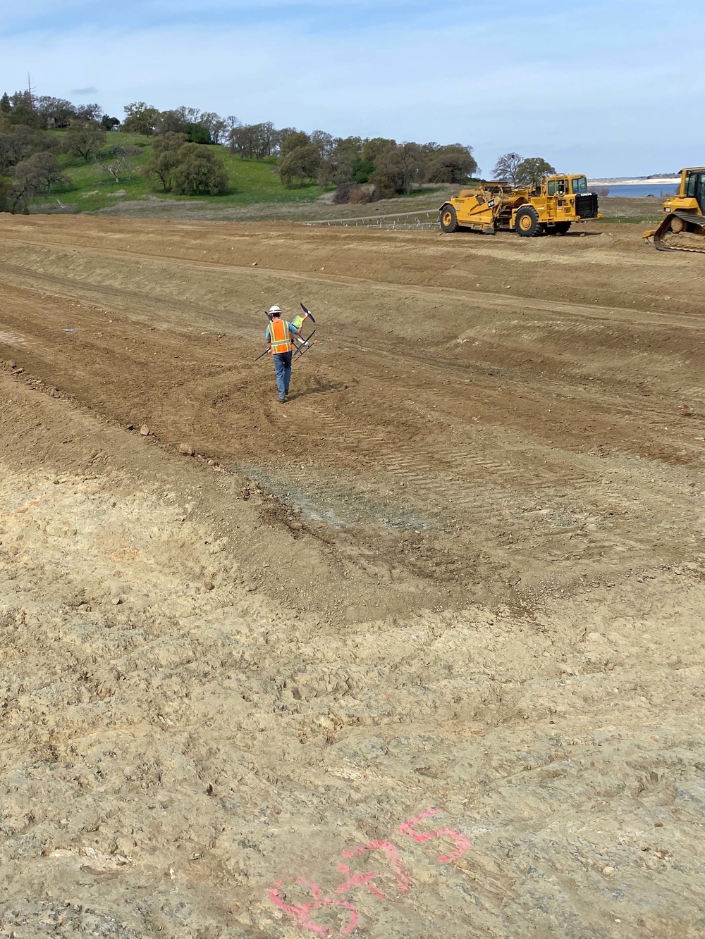 An active earthwork construction site showing a large graded dirt area near a body of water. A worker in high-visibility safety gear and hard hat is walking across the compacted soil carrying survey or marking equipment. Two large yellow Caterpillar heavy machines are visible in the background, with a green, tree-covered hillside beyond.