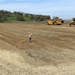 An active earthwork construction site showing a large graded dirt area near a body of water. A worker in high-visibility safety gear and hard hat is walking across the compacted soil carrying survey or marking equipment. Two large yellow Caterpillar heavy machines are visible in the background, with a green, tree-covered hillside beyond.