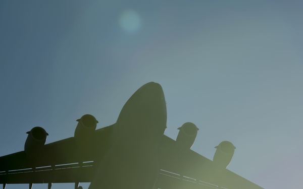 A large military transport aircraft is photographed from below against a bright blue sky, rendered almost entirely as a dark silhouette due to backlighting from the sun. The aircraft appears to be a large four-engine military transport, consistent with types such as the C-17 Globemaster III or similar heavy-lift aircraft. Lens flare and solar glare are prominently visible, significantly affecting image exposure.