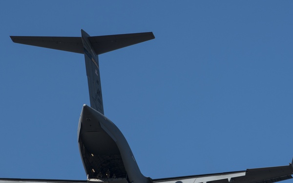 A large military transport aircraft is photographed from below against a clear blue sky. The aircraft appears to be a C-17 Globemaster III, viewed from its rear underside, with its cargo ramp visibly open during flight. The image is taken at an upward angle, silhouetting the aircraft against bright daylight.