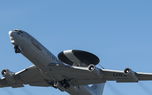 A U.S. Air Force E-3 Sentry AWACS (Airborne Warning and Control System) aircraft is captured in flight against a clear blue sky. The aircraft is identifiable by its distinctive rotating radar dome (rotodome) mounted above the fuselage and its four turbofan engines. Markings visible include 'U.S. AIR FORCE' on the fuselage and 'USA' on the tail section, with 'AK' tail code indicating Alaska-based assignment.