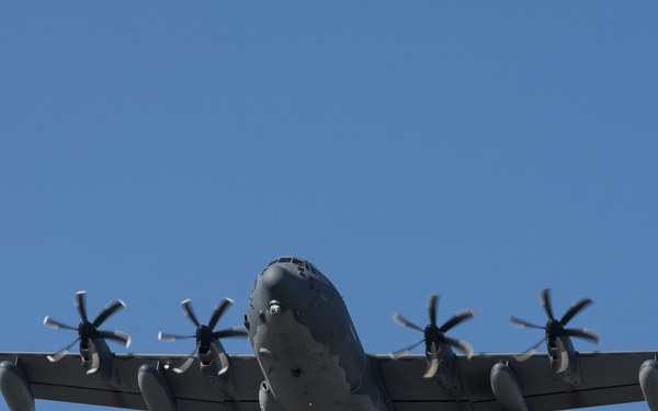 A military turboprop transport aircraft, consistent with a C-130 Hercules variant, is photographed from below in flight against a clear blue sky. The aircraft is painted in a dark gray/gunship gray color scheme, suggesting a special operations or gunship variant. All four turboprop engines are visibly spinning with multi-blade propellers engaged.