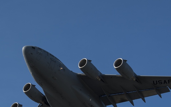 A United States Air Force (USAF) C-17 Globemaster III military cargo aircraft is photographed in flight against a clear blue sky. The aircraft is viewed from below at an angle, revealing its underside and four turbofan engines. Markings indicate it is associated with the Alaska Air Guard unit.