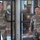 Two U.S. Army soldiers in OCP (Operational Camouflage Pattern) uniforms are standing inside what appears to be a military office or headquarters building. The soldier on the left has the name tape 'TUTT' and the soldier on the right has the name tape 'HUYNH', both wearing U.S. Army branch tapes. A display case containing plaques, awards, and memorabilia is visible in the background.