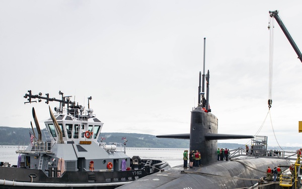 A large military submarine is docked at a naval pier, assisted by a harbor tugboat designated YT-802. Personnel in various colored vests are visible on the submarine's deck and pier, suggesting an arrival or departure operation. A yellow crane structure is positioned on the right side of the pier, likely for loading or maintenance purposes.