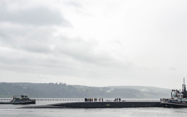 A large military submarine is shown on the surface of a calm waterway, surrounded by escort vessels and personnel visible on its deck. The scene appears to be a port arrival or departure operation, with tugboats or patrol craft flanking the submarine. The setting features a forested hillside in the background under overcast skies, consistent with a Pacific Northwest naval installation.
