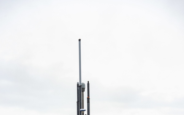A large military submarine is shown surfaced and docked or maneuvering in a calm waterway surrounded by forested hills under overcast skies. Personnel are visible on the deck and atop the sail (conning tower), with a small support/tugboat alongside on the port side. An American flag is visible on the sail, suggesting a U.S. Navy vessel, consistent with a ballistic missile or fast-attack submarine.
