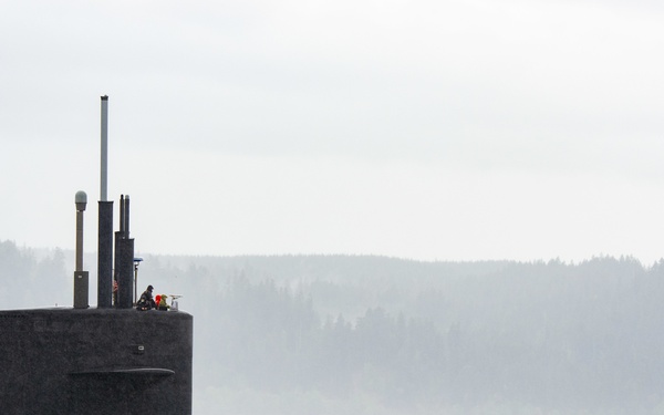 A large dark-hulled submarine is visible on the water surface, accompanied by a small harbor tugboat marked 'YT-802' on its port side. The scene appears to be a naval base or harbor operation in overcast, misty conditions with forested hills visible in the background. Personnel are visible on the submarine's sail (conning tower), and a floating barrier or boom extends across the water in the background.