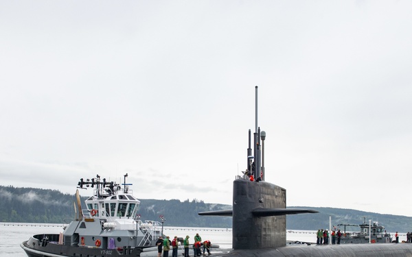 A large military submarine is surfaced on calm waters, accompanied by a tugboat bearing the designation 'YT-802'. Personnel in high-visibility vests and jackets are visible on the submarine's deck and the tugboat. The setting appears to be a Pacific Northwest waterway, indicated by the forested hillsides and overcast sky in the background.