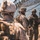 Three military personnel in desert camouflage uniforms and tactical gear are gathered near a military vehicle under a camouflage netting. One soldier in the foreground is out of focus, while two others in the background appear to be reviewing information on a tablet or clipboard. The setting suggests a field operation or training exercise in an arid environment.