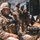 The image shows military personnel in desert camouflage uniforms operating in a field environment under camouflage netting. The foreground soldier is using a field radio handset while equipped with full tactical gear. Additional soldiers are visible in the background working near what appears to be military equipment or a vehicle.