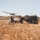 Two individuals in military-style attire are operating a fixed-wing UAV (unmanned aerial vehicle) in an open dry grassland field. One person is holding the UAV overhead, likely preparing for a hand-launch, while the other stands near camouflage-netted military vehicles. The setting appears to be a training or field operation exercise in an arid, rural environment.
