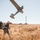 A military personnel in camouflage uniform is hand-launching a fixed-wing unmanned aerial vehicle (UAV) in an open dry grassland field. A camouflage-netted military vehicle is visible in the background along with utility poles and power lines. The scene appears to be a field training or operational exercise.