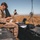 Two military personnel in desert camouflage uniforms are operating in an open field with dry grass. The foreground soldier is seated on hard-shell equipment cases handling what appears to be a small UAV or drone component, with a ruggedized laptop and communications equipment nearby. A second soldier stands in the background, appearing to hold a controller or tablet device.