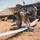 Two military personnel in desert camouflage uniforms are operating a fixed-wing unmanned aerial vehicle (UAV) in an open field with dry grass. One soldier is bent over preparing or inspecting the UAV on the ground, while a second stands near a camouflage-netted field shelter with equipment cases. The setting appears to be a forward operating position or field exercise location.