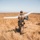 A person in military-style camouflage gear is carrying a fixed-wing UAV (unmanned aerial vehicle) through a dry, golden grassland field. The individual is facing away from the camera and appears to be preparing for a hand-launch of the small drone. The setting appears to be an open, arid terrain under a clear sky, consistent with a training or operational environment.