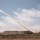 A rocket or missile is being launched from a ground-based vehicle in an open, arid field. A long white smoke trail extends diagonally from the launch vehicle up toward the upper left of the frame. The setting appears to be a military test range or exercise area, with earthen berms visible in the background.