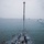 USS Gladiator (MCM 11) A view from an elevated position looking down the bow of a gray naval vessel underway on overcast, choppy waters. Several crew members wearing safety helmets and vests are working on the foredeck with lines and mooring equipment. Multiple vessels, likely tugboats, are visible in the background suggesting a port departure or arrival operation.