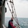 Damage Controlman Jason Anderson inspects a rigid-hull inflatable USS Gladiator (MCM 11). A masked crew member wearing red coveralls and a blue vest is working on the deck of a sailing vessel, attending to equipment on or near an inflatable rigid-hull boat (RIB/RHIB). Multiple hemp or natural-fiber rigging ropes and a metal davit hook are visible overhead, suggesting the RIB is being prepared for launch or recovery. The scene takes place at sea under an overcast sky with calm open water visible in the background.