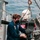 USS Gladiator (MCM 11) A young person wearing a dark blue coverall uniform and black face mask is seated in a rigid inflatable boat (RIB), working on or inspecting the helm/steering console. The boat is suspended or docked alongside a larger grey naval or military vessel, with rigging and a crane hook visible overhead. An Evinrude outboard motor is visible at the stern of the RIB.