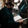 Chief Electricians Mate Maritza Hettinger USS Gladiator (MCM 11) A person wearing a teal/dark green uniform and black face mask is reviewing documents in a large binder in what appears to be a cramped, industrial workspace. The environment is cluttered with equipment, binders, and supplies on shelves, suggesting a technical or operational setting such as a military vessel or industrial facility. The lighting is dim with a single overhead light source illuminating the workspace.