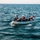 USS Gladiator (MCM 11) retrieve SeaFox from the ocean Three individuals are aboard a gray inflatable rigid hull inflatable boat (RHIB) on open water. The crew members are wearing safety gear including helmets and life vests, and an American flag is visible mounted on the vessel. The boat appears to be underway, with visible wake and choppy sea conditions.