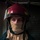 Engineman 3rd Class Alexander Scott prepares for a fire drill A firefighter is photographed in a close-up portrait wearing full protective gear including a red helmet with a clear visor and yellow reflective markings, and a tan/beige fire-resistant hood covering the lower face and neck. The subject appears to be indoors in a dark industrial or structural environment. The image has a dramatic, moody lighting style with the subject's eyes being the primary focal point.