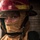 Engineman 3rd Class Alexander Scott prepares for a fire A close-up portrait of a firefighter wearing full protective gear, including a red helmet with a raised clear face shield and a tan/yellow fire-resistant hood covering the lower face. The subject is looking directly at the camera with a composed expression. The background is dark and out of focus, suggesting an indoor or low-light environment.