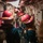 Fire Drill Aboard USS Gladiator (MCM 11) A group of personnel wearing red firefighting helmets and protective gear are moving through a narrow corridor aboard what appears to be a naval vessel. They are equipped with breathing apparatus (SCBA tanks) and fire hoses, suggesting a damage control or firefighting drill. The confined passageway is lined with pipes, cables, and electrical panels typical of a ship's interior.